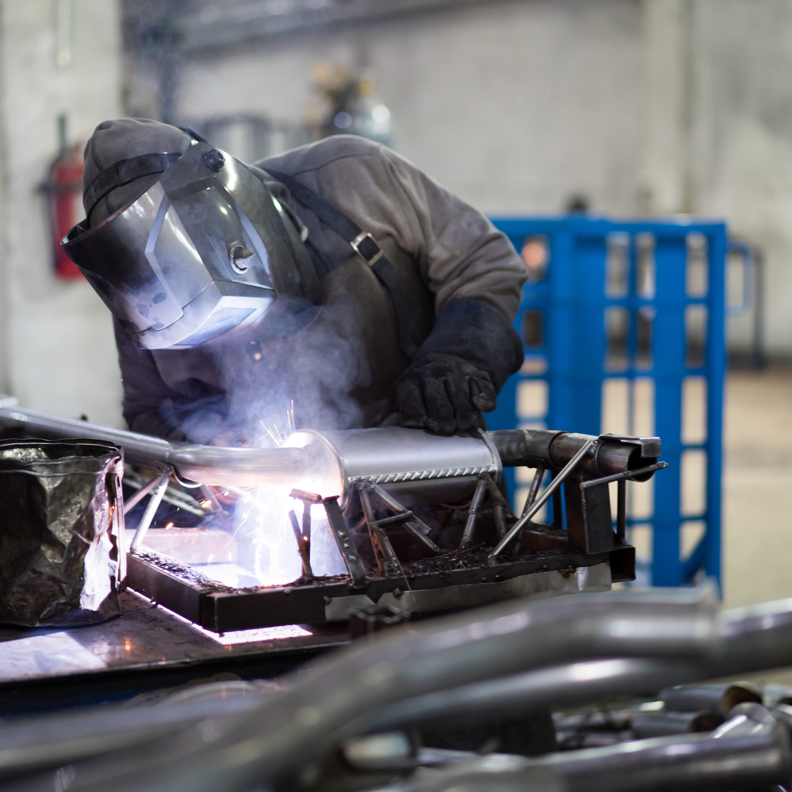 view of a worker wearing protective gear, with mask, leather apron and gloves, welding together pieces of an exhaust pipe, with several other pipes around, on the work table, and metal blue frames in the background, in an industrial setting